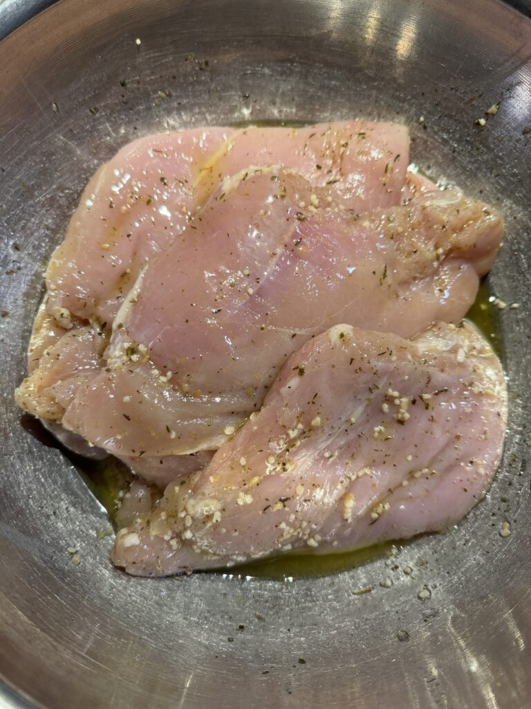 Raw chicken breasts coated in olive oil marinade resting in a bowl before grilling